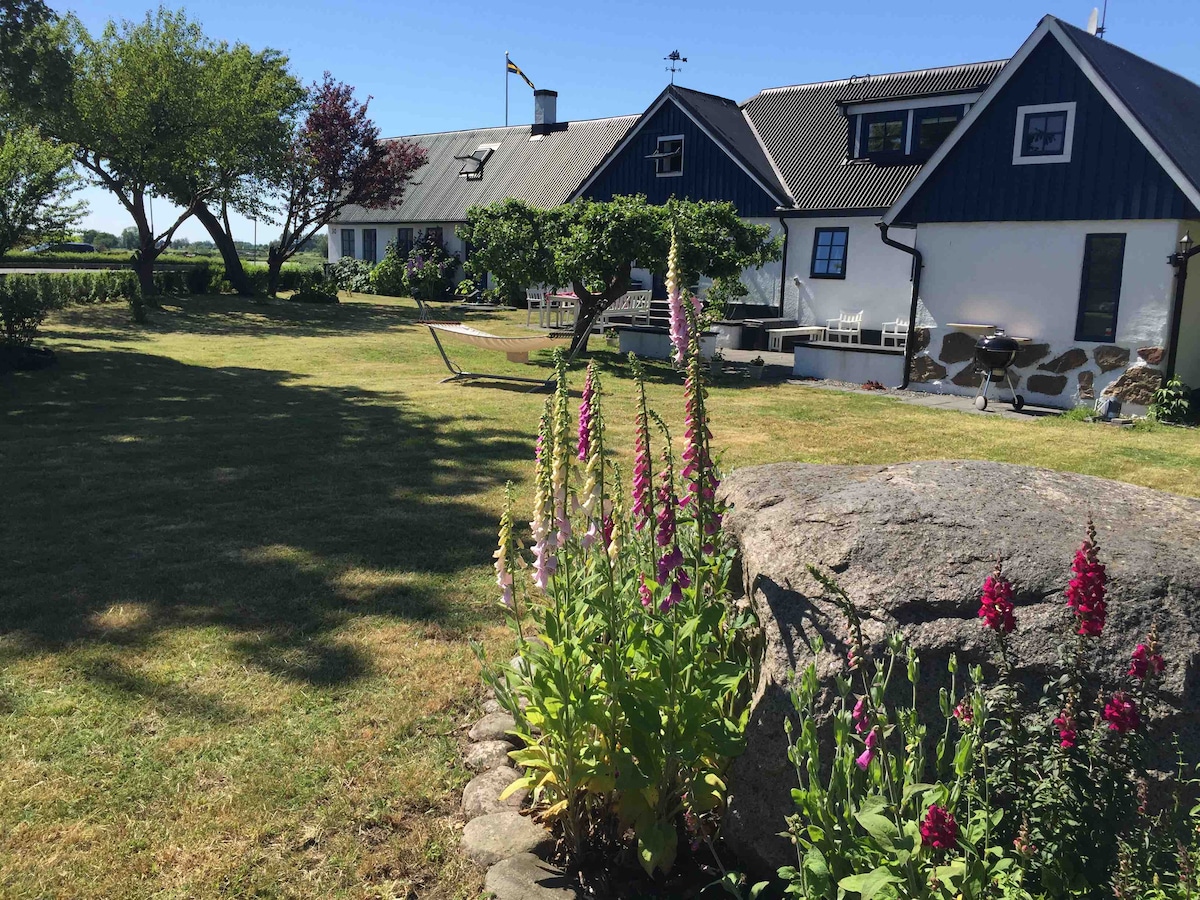 A spacious garden is highlighted by lush, manicured grass and colorful flower beds featuring tall blossoms. The charming farm house is visible in the background, showcasing a combination of traditional and modern architectural elements. Trees provide shade while a hammock invites relaxation.