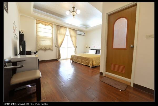 A spacious room features a queen size bed with a decorative headboard. Natural light enters through a window with soft drapes, enhancing the warm tones of the wooden flooring. A door leads to an exterior area, while a small table and chair are positioned nearby.