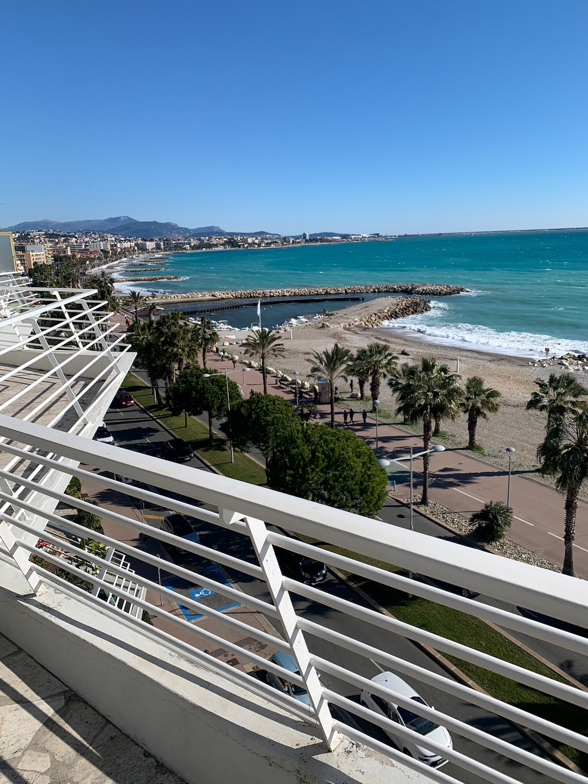 A broad view encompasses the shoreline and sparkling blue waters, framed by palm trees and sandy beach. The coastline is marked by a gently curving promenade, while a small harbor is visible in the distance under a clear blue sky.
