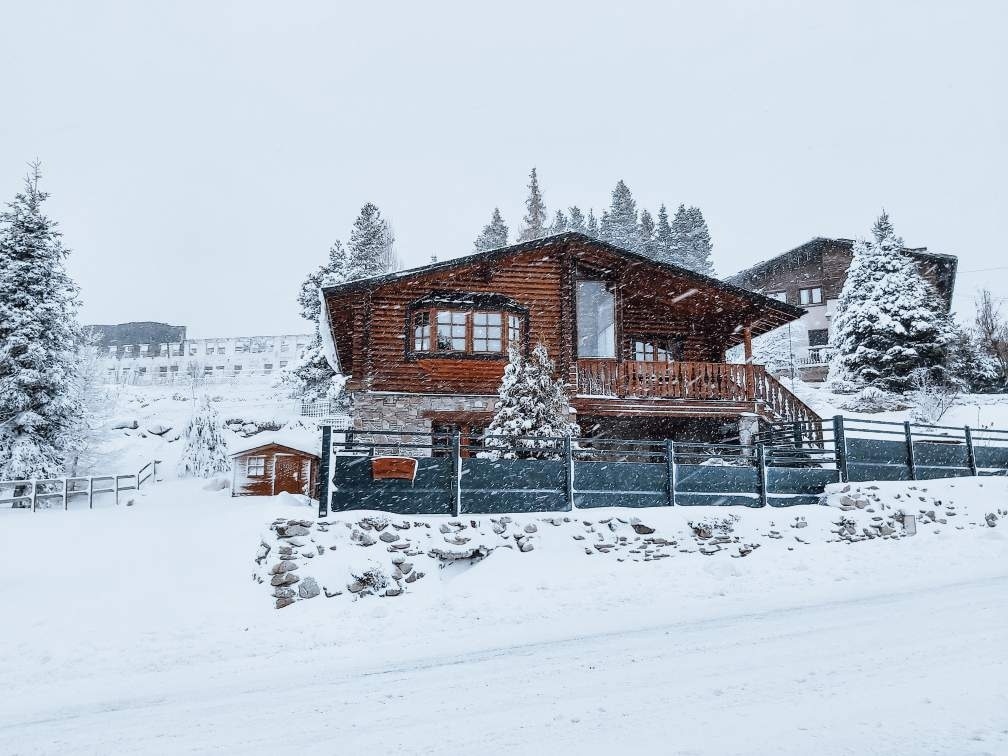 A wooden chalet is depicted in a snowy landscape, surrounded by trees and a fenced area. Snow covers the ground and rooftops, creating a serene winter scene. The structure features a spacious balcony and large windows, allowing for natural light to enter.