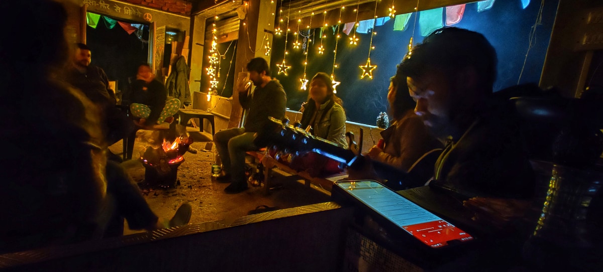 A cozy gathering around a crackling bonfire is shown, with people seated on wooden benches. Twinkling string lights and colorful bunting create an inviting atmosphere in the dimly lit space. A guitar is resting on a table, enhancing the serene evening vibe.