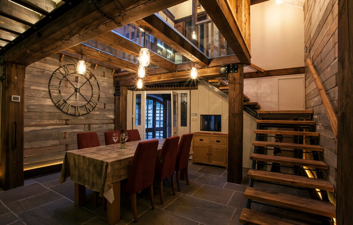A rustic dining area features a large wooden table surrounded by red upholstered chairs. Exposed wooden beams and a wall-mounted clock complement the warm lighting, while a staircase leads to the upper level. A glass floor floods the space with natural light.