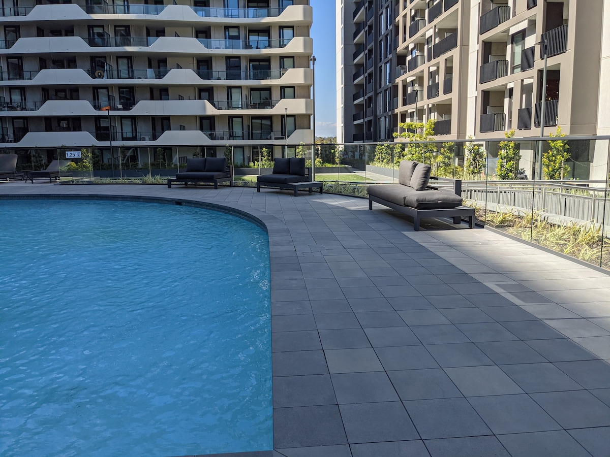 A spacious outdoor area features a calming swimming pool surrounded by modern loungers. The well-maintained patio is made of large tiles, with green landscaping visible in the background, creating a relaxing ambiance for guests.