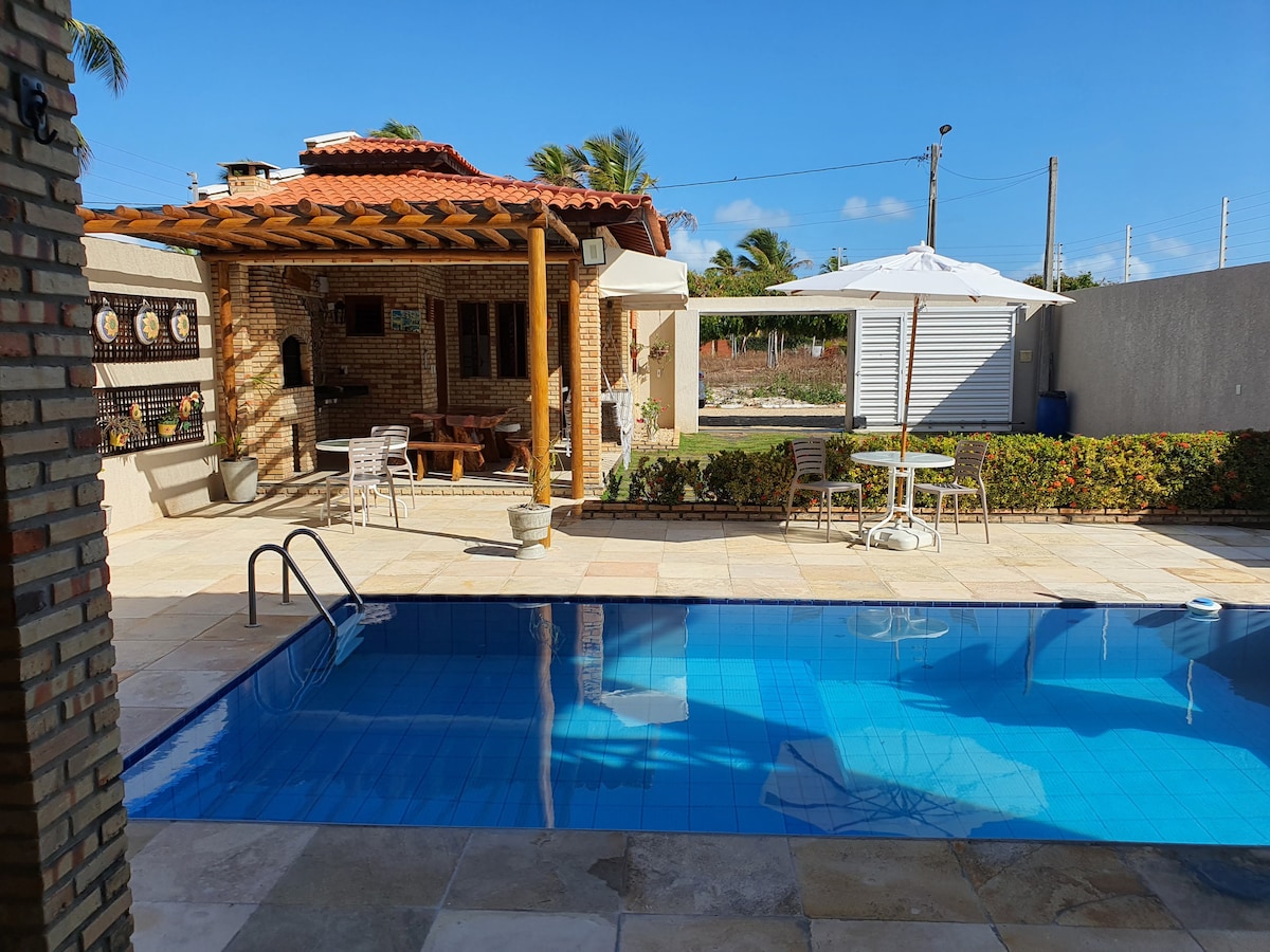 An outdoor area features a swimming pool with steps leading in, surrounded by tiled flooring. A covered seating area is visible, consisting of wooden furniture under a thatched roof. Sun loungers and a white umbrella are arranged nearby, with greenery lining the perimeter.