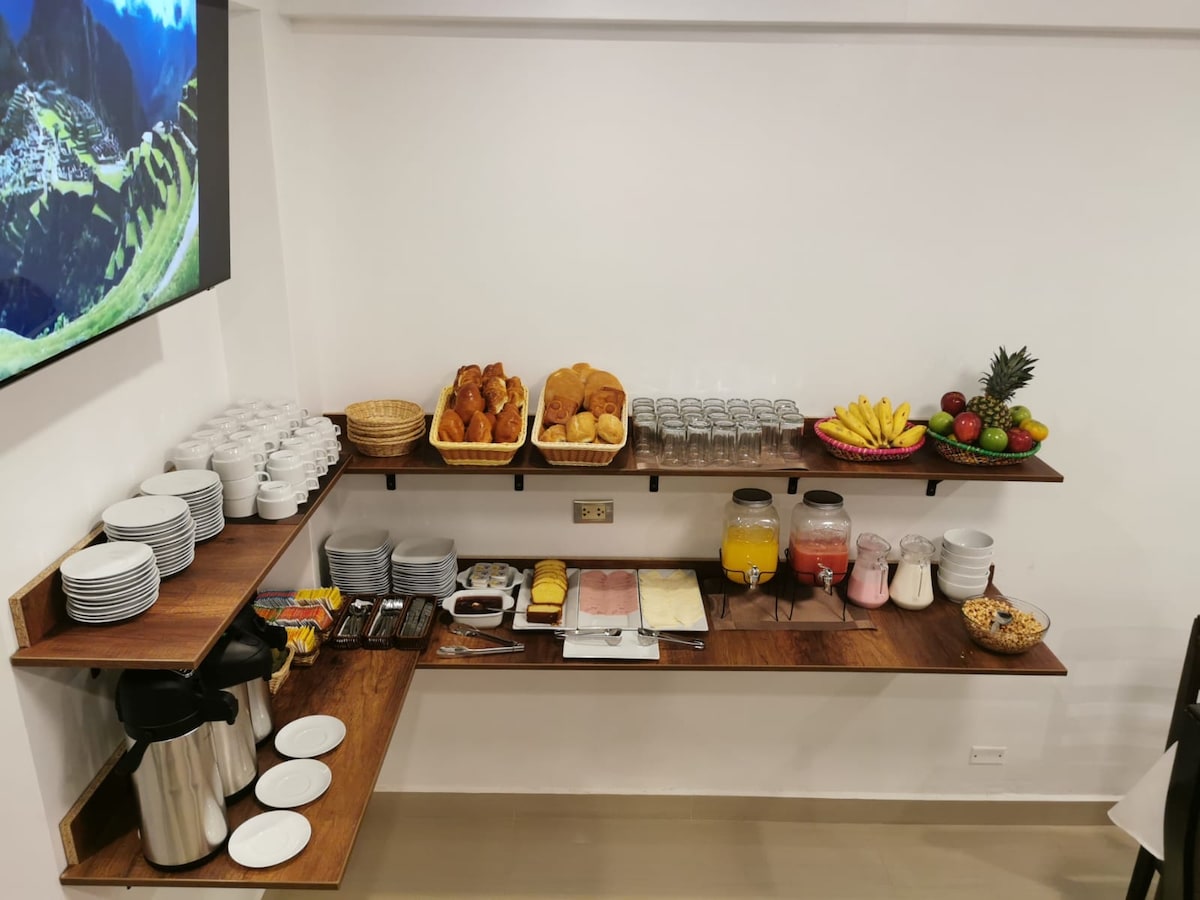 A buffet area is presented with two wooden shelves showcasing a variety of breakfast items. Freshly baked bread and pastries are arranged next to fruit bowls. Beverages, including juice and milk, are stored in clear containers. Plates, cups, and glasses are neatly organized for guest use.