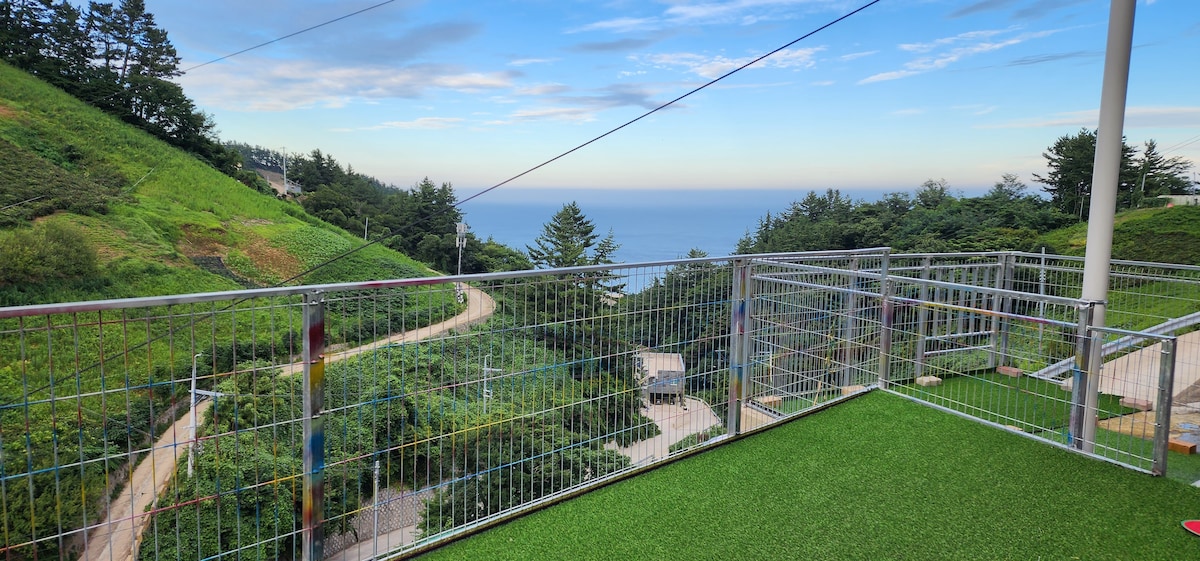 An outdoor deck overlooks a lush landscape and the ocean in the distance. A winding path can be seen, surrounded by greenery. Fencing surrounds the deck area, enhancing the open view and providing a sense of safety.