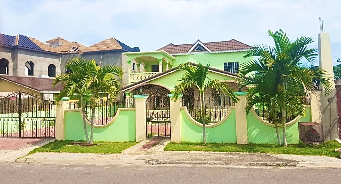 The exterior of a spacious green property is displayed, featuring a decorative wrought iron gate and lush palm trees. The well-maintained garden area is visible in front of the home, with a two-story structure in the background displaying a classic architectural style.