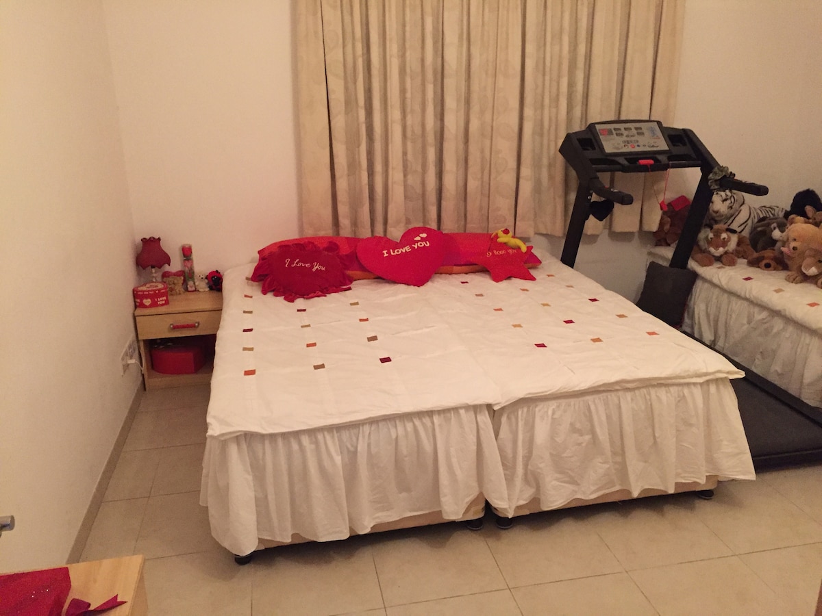 A spacious bed is covered with a white bedspread featuring colorful squares. Red pillows with playful phrases adorn the bed. Nearby, a treadmill is positioned next to a bedside table containing decorative items. Soft curtains frame a window, allowing natural light to fill the room.