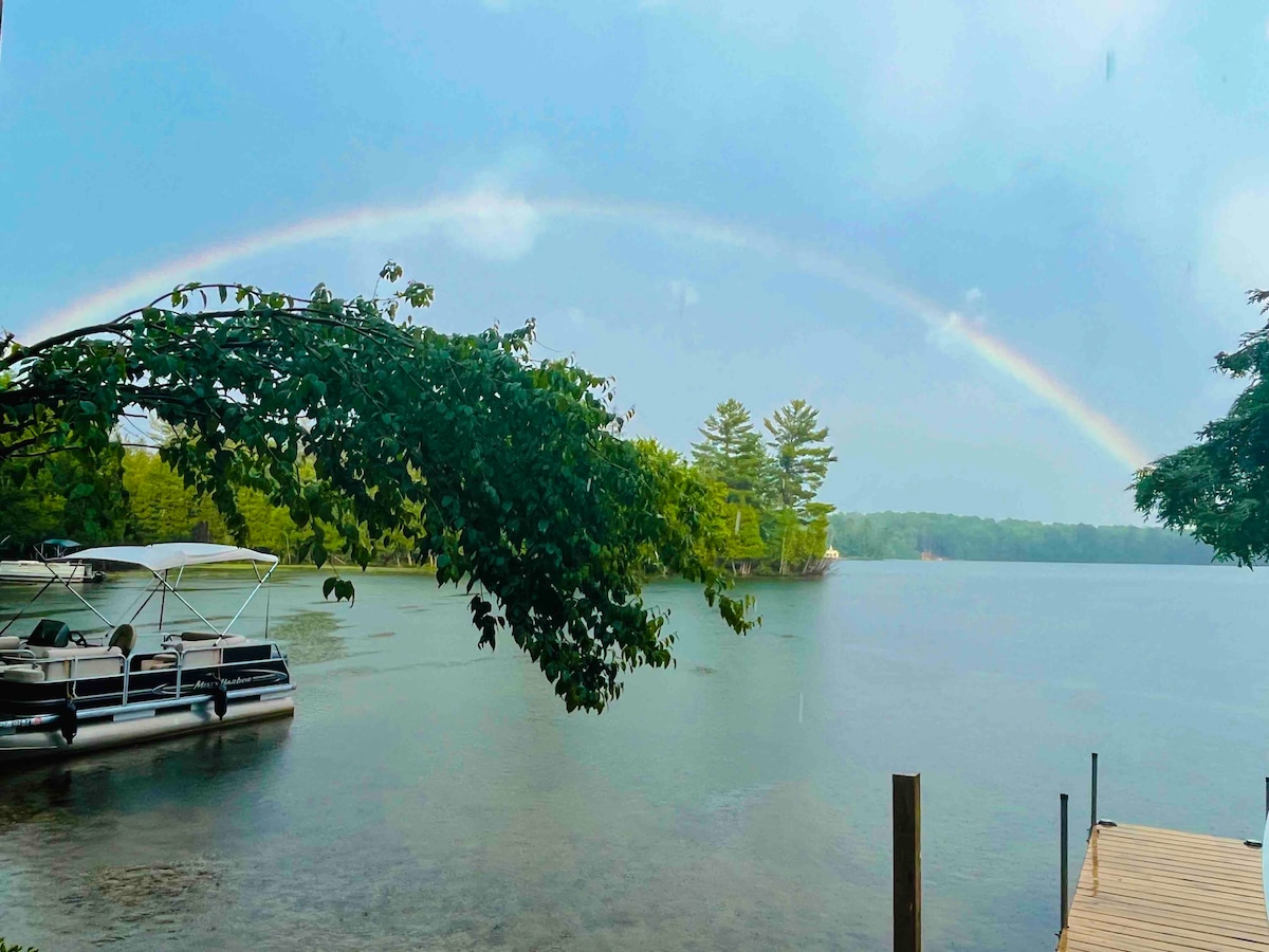 A serene lake is viewed under a rainbow arching across the sky. A dock extends into the water, alongside a pontoon boat anchored nearby. Lush green trees frame the scene, enhancing the tranquil atmosphere of the surroundings.