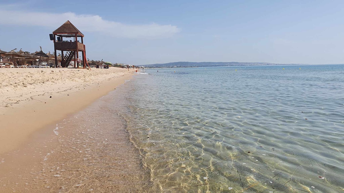 A sandy beach is visible with gentle waves lapping at the shore. The clear water allows for visibility of the sandy bottom. In the distance, beachgoers can be seen relaxing under a bright sky, while a lifeguard tower stands on the shore.