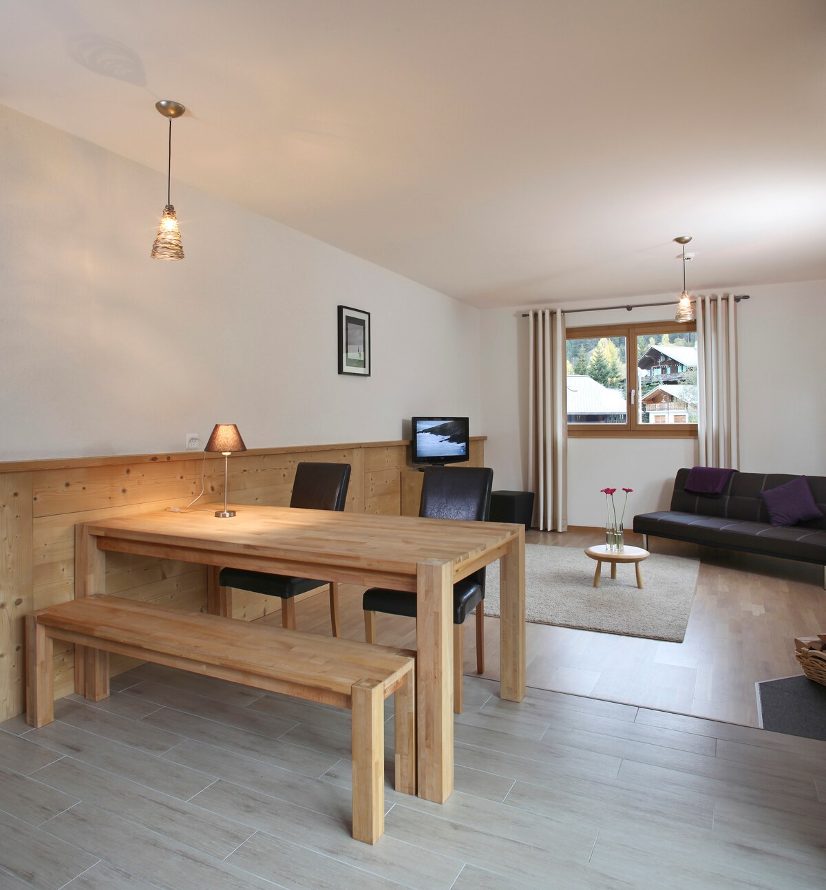 A modern dining area features a wooden table accompanied by a long bench and two black chairs. A small lamp illuminates the table. The adjacent seating area includes a dark sofa, a rug on a light floor, and a window with sheer curtains, revealing a view outside.