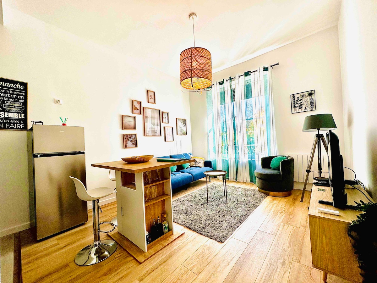 The living area features an open layout, with a cozy blue sofa and a modern armchair. A light wooden dining island serves as a central feature, complemented by a bar stool. Large windows dressed in sheer curtains allow natural light to illuminate the space.