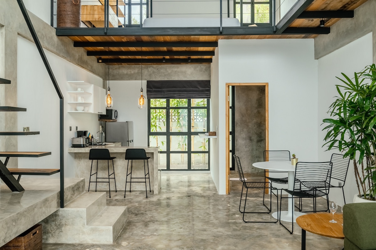 A spacious and open living area is featured, showcasing a modern aesthetic with concrete walls and a blend of natural wood elements. A well-equipped kitchen is visible, along with a dining area featuring a round table and four chairs. Large sliding doors allow natural light to fill the space.