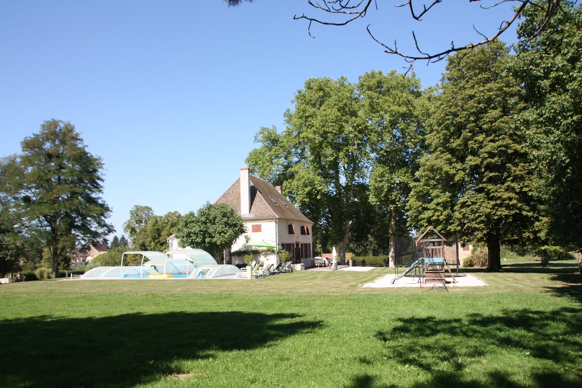 The elegant 19th-century house is framed by lush green lawns and tall trees, providing shade. A covered swimming pool is visible nearby, alongside a playground featuring a swing set and sand area, creating a family-friendly atmosphere in a serene outdoor setting.