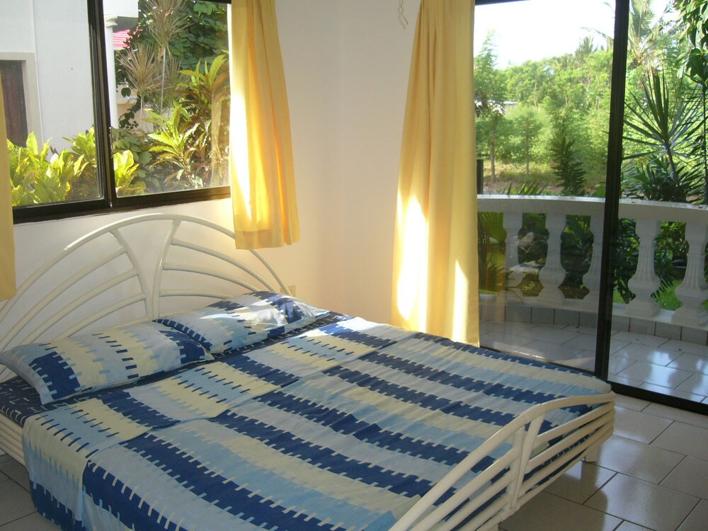 A bright sleeping area is featured, with a spacious white bed dressed in blue and white patterned linens. Natural light filters through large windows draped with yellow curtains, revealing a lush garden outside.