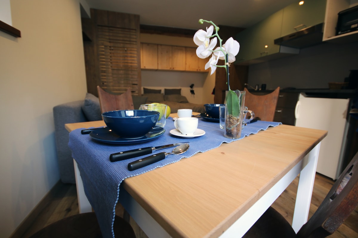 A dining table is set for a meal, featuring blue plates, a coffee cup, and cutlery arranged neatly. A small vase with white flowers adds a decorative touch. In the background, a cozy living area is visible, complementing the inviting dining space.
