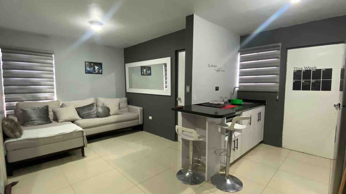 A modern living space is presented with a light-colored sectional sofa against a gray wall, complemented by decorative pillows. A kitchen bar with two stools is visible, along with sleek cabinetry and a door featuring a simple design. Natural light is provided by window coverings.