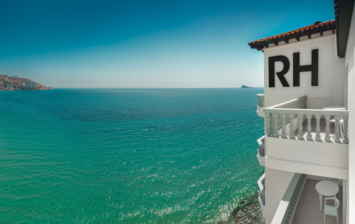 A coastal view captures the shimmering turquoise waters of the Mediterranean Sea, framed by the white balcony of the hotel. The prominent hotel logo, RH, is displayed, emphasizing the destination's inviting atmosphere. A distant coastline appears along the horizon, blending seamlessly with the sky.