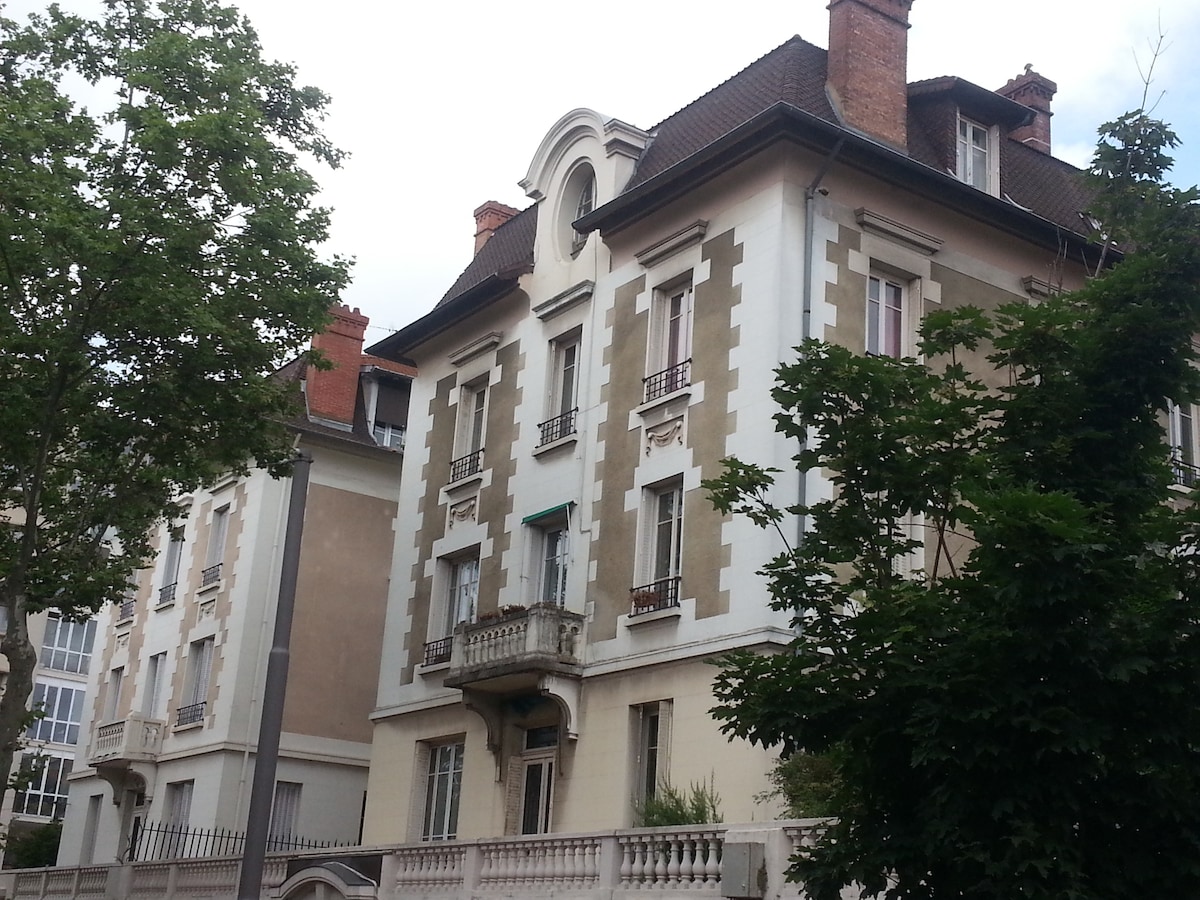 A historic building with a classic architectural style is surrounded by greenery. The three-story structure features a blend of beige and white facades, with decorative elements above the windows. Balconies create visual interest, while trees frame the exterior, contributing to a serene environment.