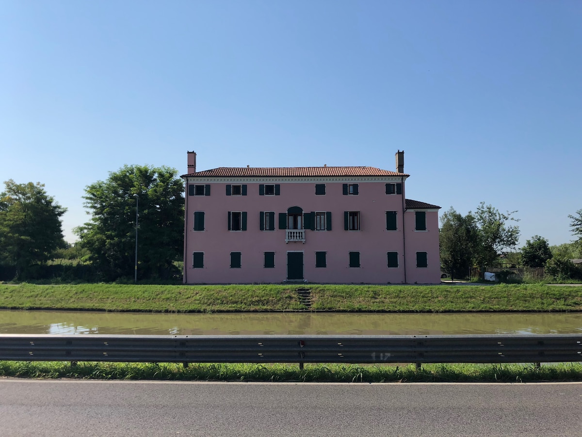 A historic Venetian hunting lodge, presented in a soft pink hue, is situated alongside a peaceful waterway. The structure features multiple windows with green shutters and a central balcony, framed by lush greenery on either side.