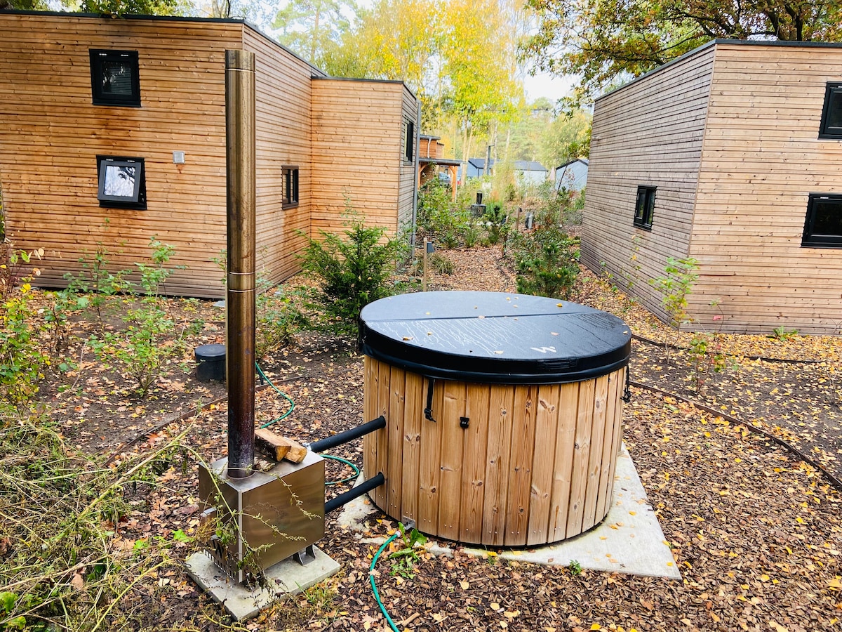 A wooden hot tub is situated on a concrete base surrounded by foliage, contributing to the natural setting. A metal chimney is attached to the tub, indicating a heating system. The nearby cabins, designed with wood exteriors and black window frames, create a cozy atmosphere.