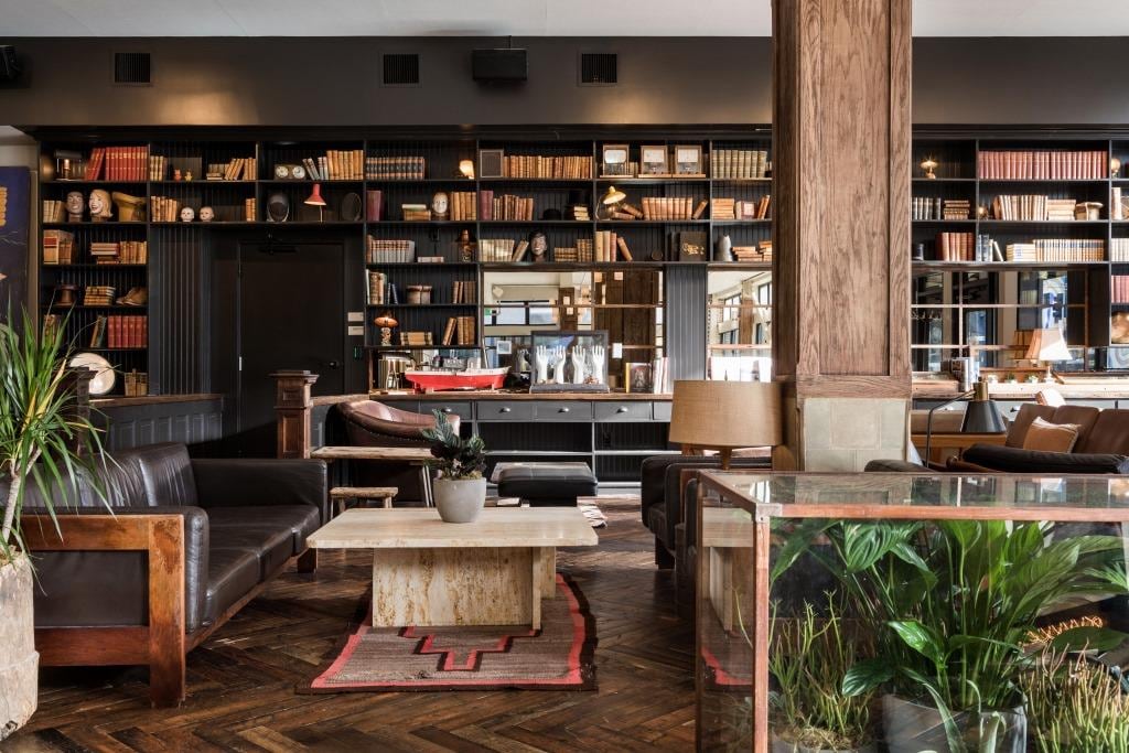 The inviting lounge area is furnished with dark leather sofas and a warm wooden coffee table. A wall of bookshelves filled with books and decorative items fosters a cozy ambiance. Green plants add a touch of nature, complementing the rich textures of the space.