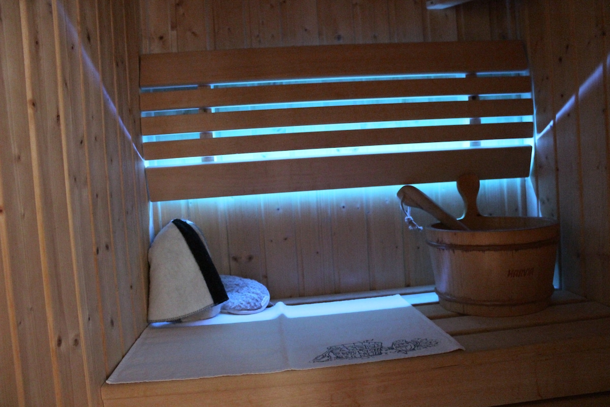 A soothing sauna interior is highlighted, featuring light wood paneling and a slatted wooden bench. A light source softly illuminates the space, enhancing the serene atmosphere. A wooden bucket and towel are placed for convenience, creating a space for relaxation.