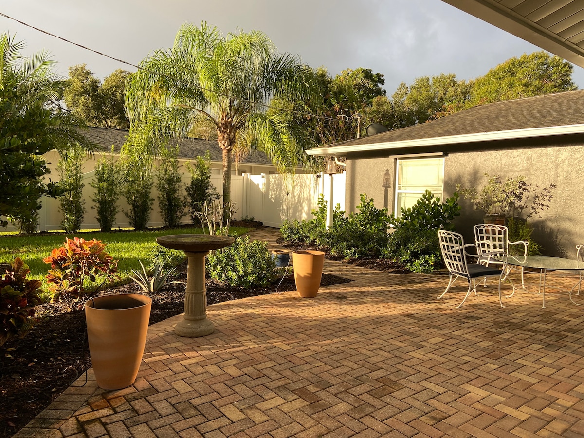 A private backyard space features a stone-paved area surrounded by lush greenery and landscaping. Two outdoor chairs are positioned adjacent to a small stone table, complemented by terracotta planters. A birdbath adds a charming focal point amidst the serene environment.