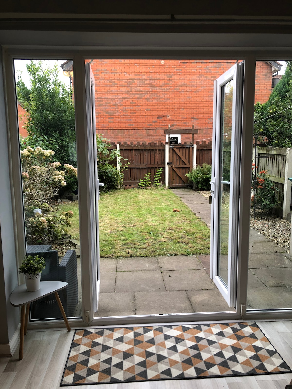 A backyard area is visible through open glass doors. The space is defined by well-maintained grass and a mix of shrubs and trees along the perimeter. A small patio is positioned just outside the doors, with a decorative mat and a potted plant adding visual interest.