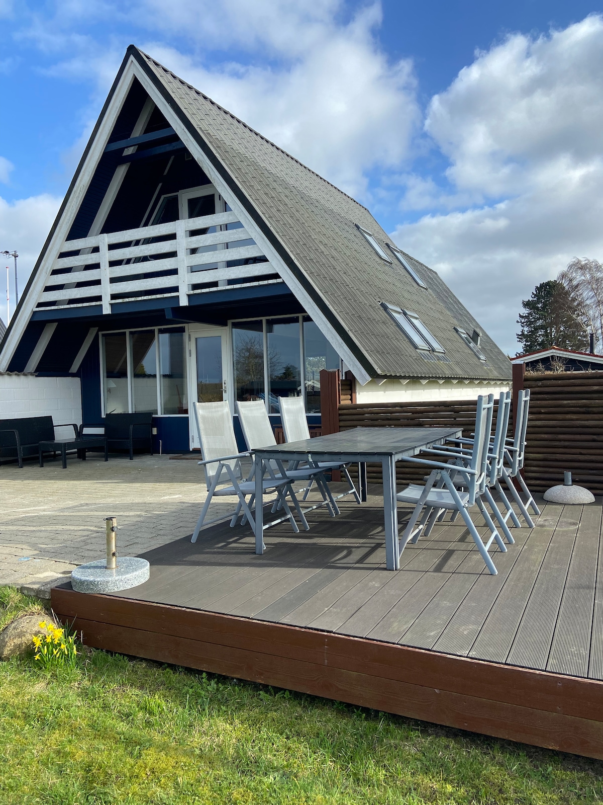 A two-story A-frame house is positioned in a tranquil outdoor setting, featuring a spacious deck furnished with a large dining table and chairs. Large windows of the house allow for ample natural light, and a section of lush green grass frames the wooden deck.
