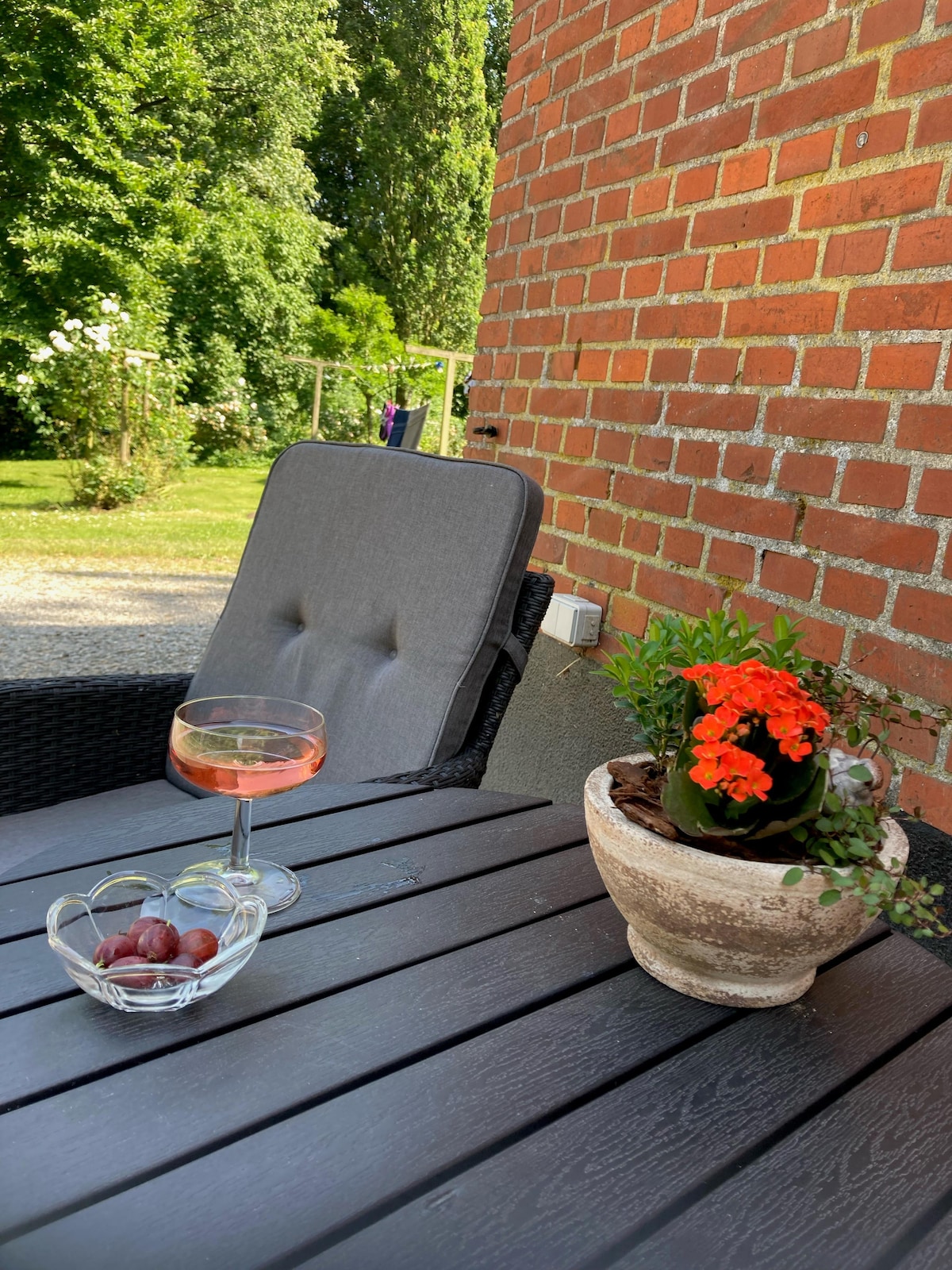 A comfortable outdoor seating area features a plush gray chair beside a dark wooden table. A glass of beverage rests on the table next to a bowl of grapes and a flower pot containing vibrant orange flowers. Lush greenery is visible in the background.
