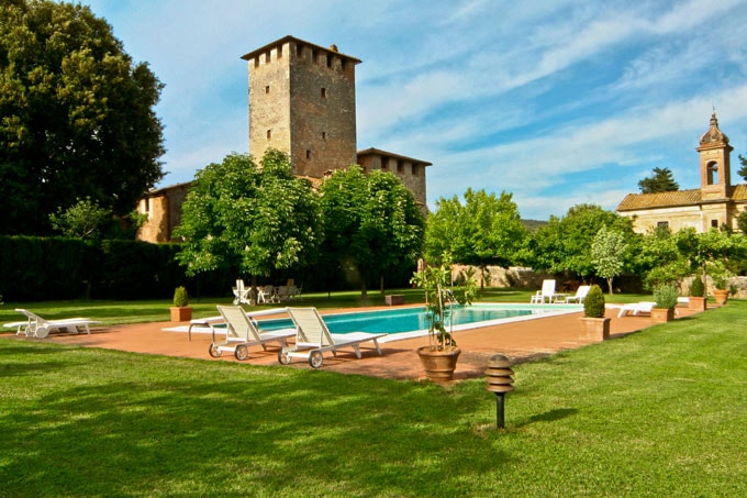 The outdoor area features a serene swimming pool surrounded by a well-maintained lawn. Lounge chairs are positioned along the poolside, while lush trees and potted plants provide natural shade. A historic stone tower is visible in the background, contributing to the peaceful ambiance.
