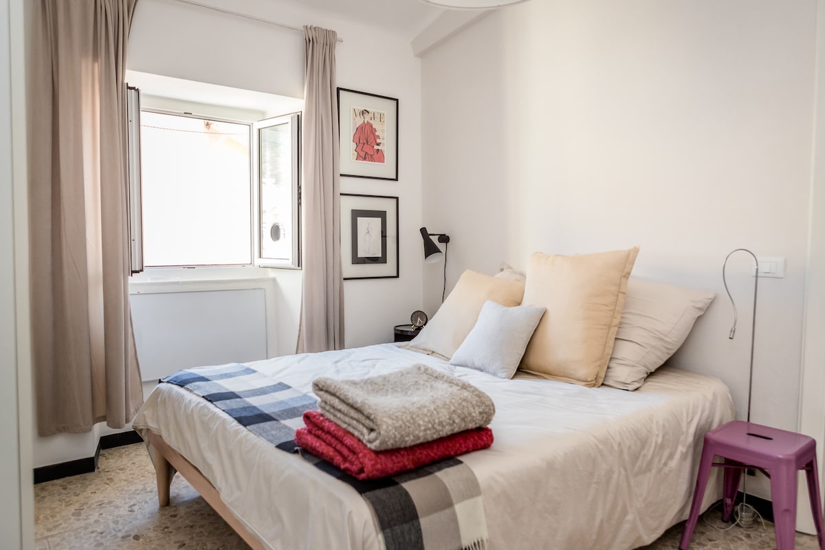 A bright bedroom is highlighted by natural light streaming through a large window. A spacious bed is adorned with a variety of pillows and neatly folded blankets. A small stool and framed artwork add character to the inviting space.