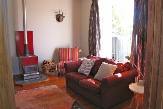 A cozy living area features a red wood stove, providing warmth, and a red couch adorned with decorative pillows. Natural light is allowed to flow in through the large window, which is framed by patterned curtains. A basket of firewood is placed nearby, enhancing the inviting space.
