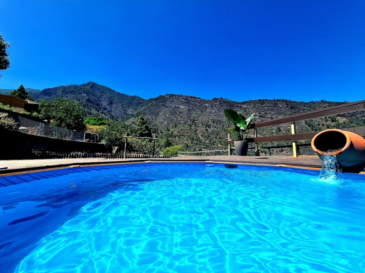 A bright blue pool is captured in the foreground, surrounded by wooden decking. Lush green mountains are visible in the distance against a clear blue sky. A potted plant adds a touch of greenery beside the pool, enhancing the natural setting.
