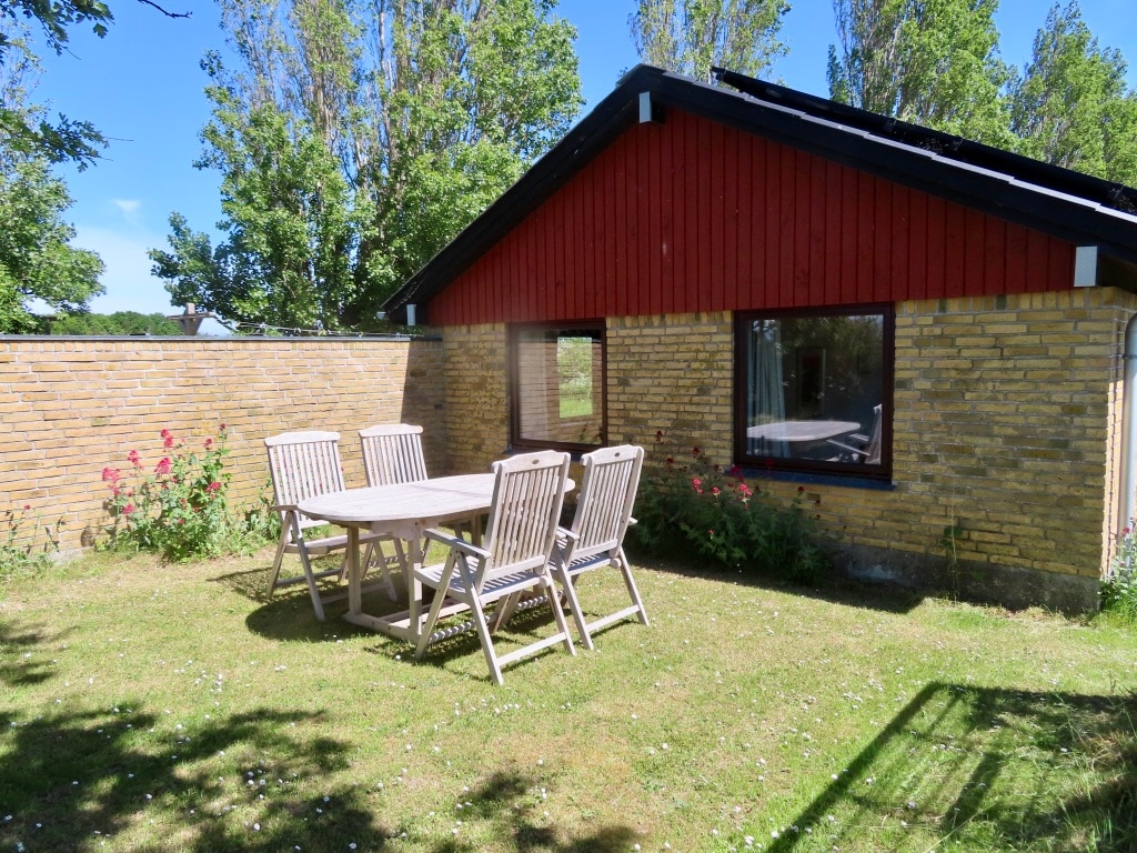 A charming outdoor space is depicted, featuring a wooden dining table surrounded by matching chairs. A well-maintained lawn is visible, dotted with flowers, and the red and yellow brick exterior of the building complements the greenery. Sunlight filters through the trees, providing a serene ambiance.