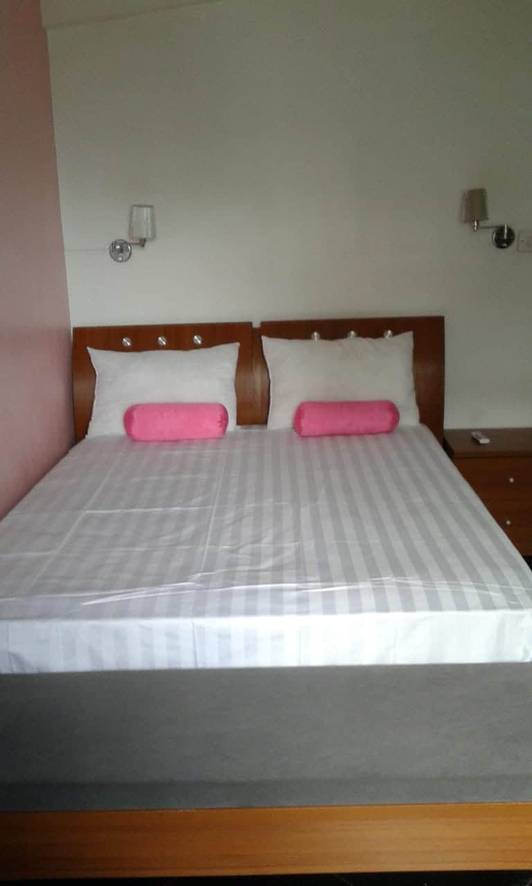 A spacious bed features a light striped covering with two white pillows and two pink accent pillows. The wooden headboard is complemented by wall-mounted lamps. Nearby, a nightstand offers additional storage space.