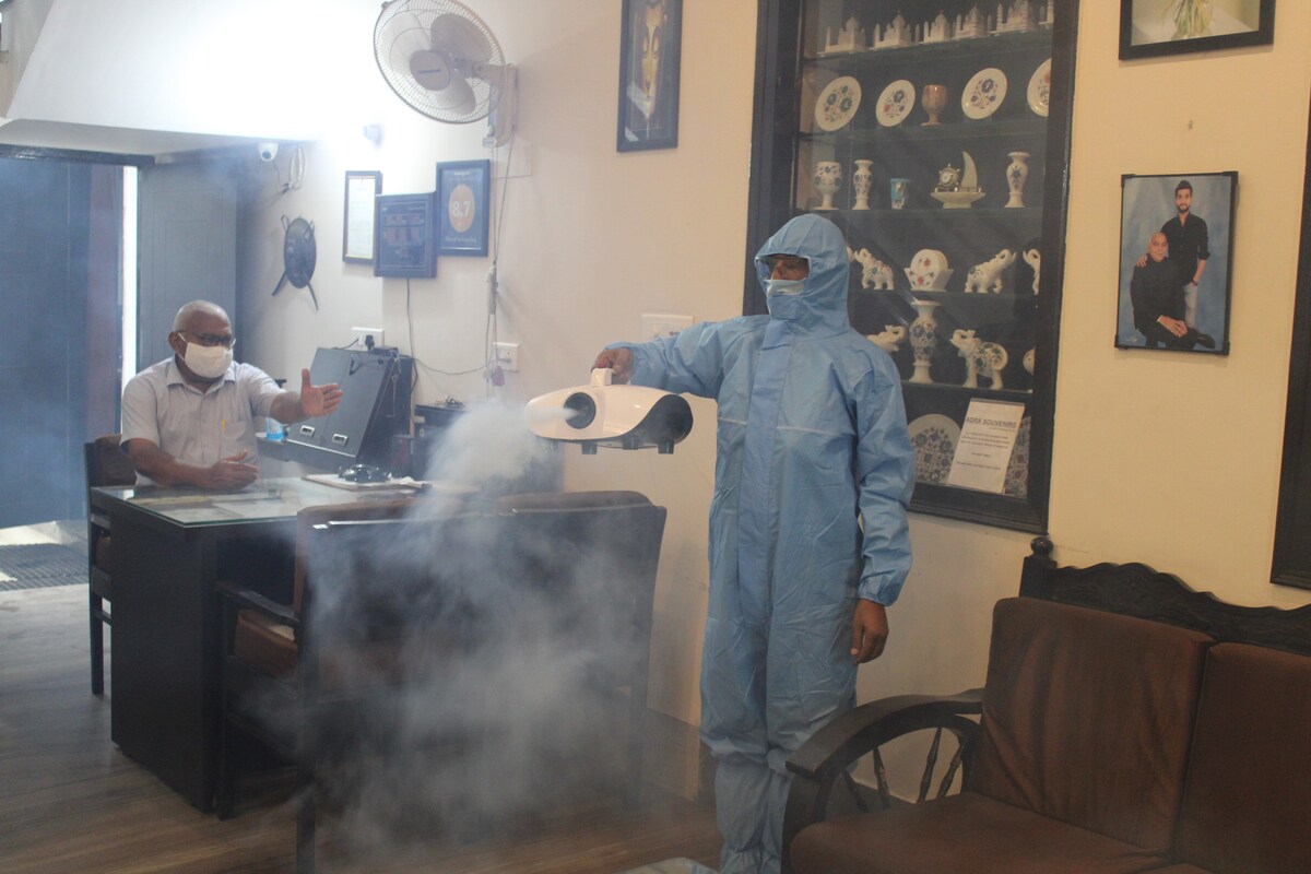 A health safety professional, dressed in protective gear, is seen sanitizing the reception area. Equipment used for disinfection is held over a surface, with a light mist visible. A staff member is seated at the reception desk, observing the process.