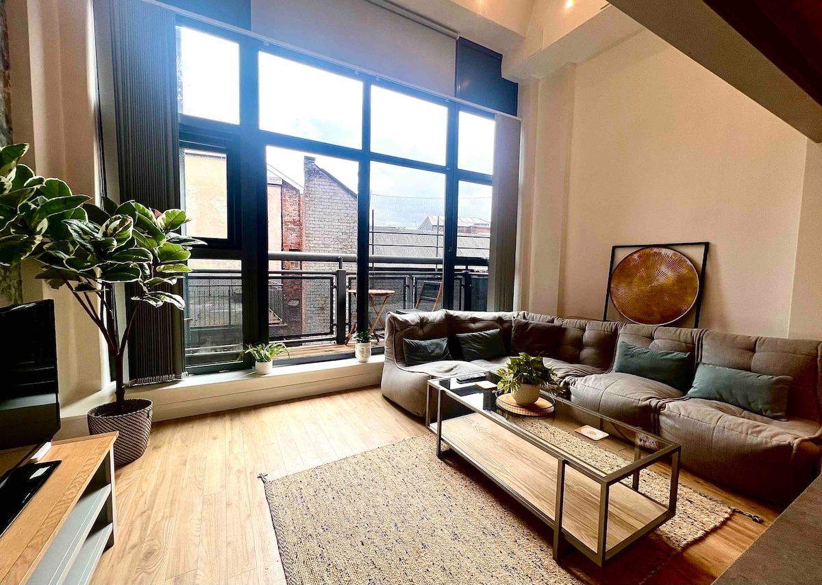 A spacious living area features large windows that allow natural light to fill the room. A sectional sofa is accompanied by a coffee table and a few decorative plants. A textured area rug lies on the wooden floor, complementing the modern decor.