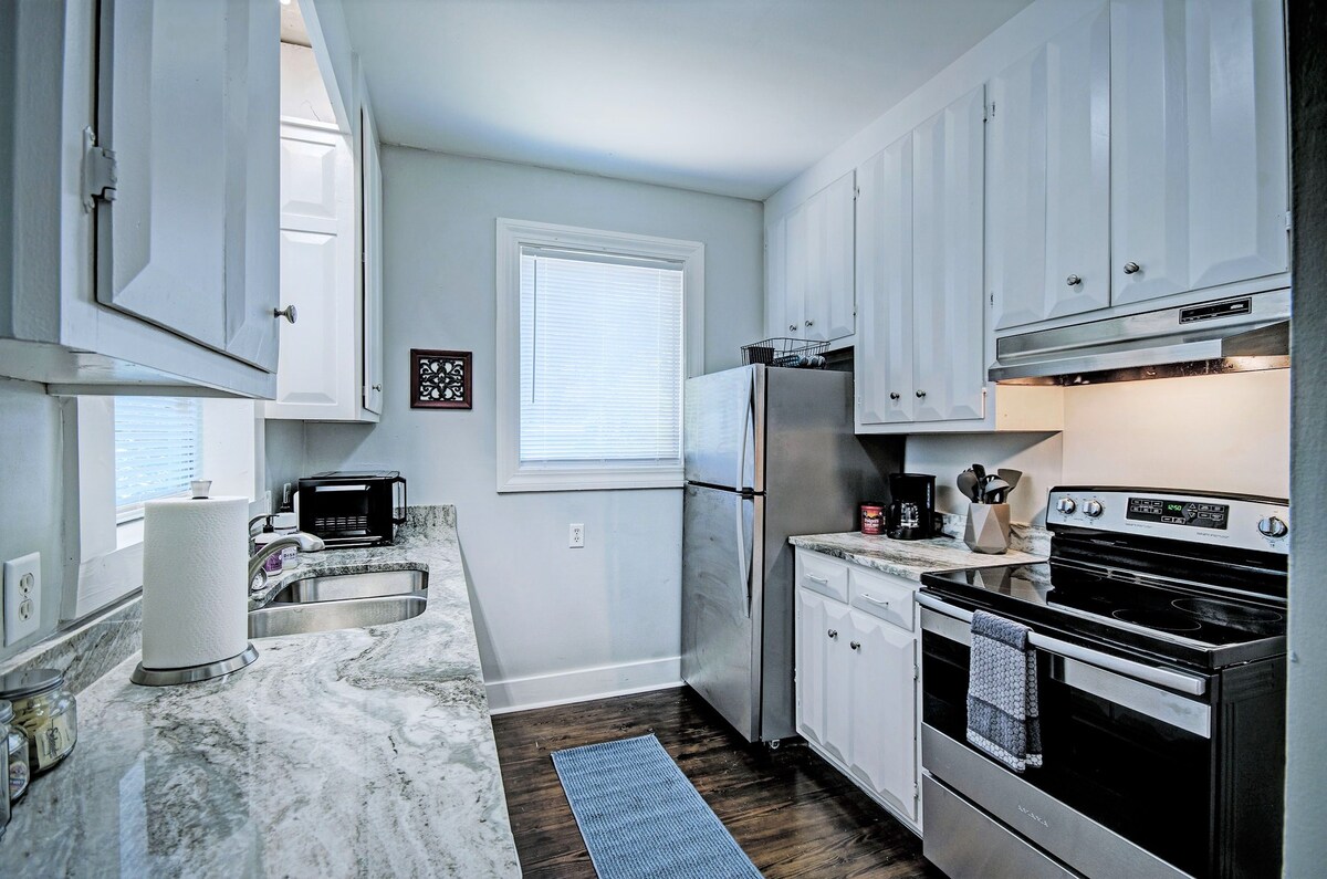 The kitchen features white cabinetry and a granite countertop, offering a modern design. Stainless steel appliances, including a refrigerator and stove, are present. Natural light enters through a window, creating a bright and functional space. A blue mat is positioned on the dark hardwood floor.