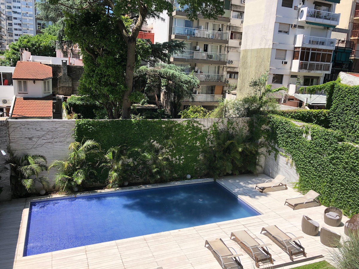 The outdoor area features a rectangular swimming pool surrounded by wooden decking. Lush greenery provides privacy and enhances the tranquil setting. Multiple lounge chairs are placed around the pool, inviting relaxation under sunlight, while a small circular seating area is visible in one corner.