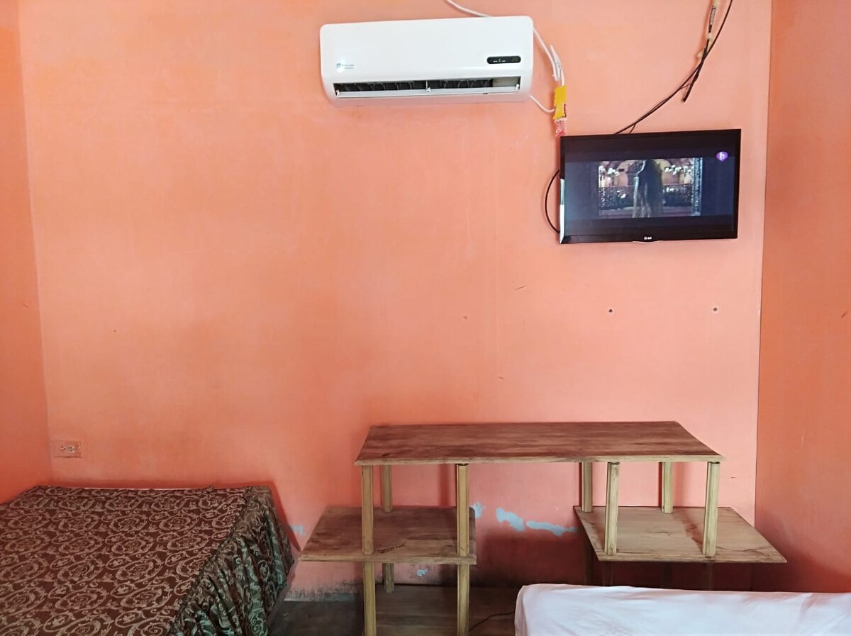 A simple room features a wall-mounted television above a wooden table with storage compartments. An air conditioning unit is installed near the ceiling. One bed is presented with a patterned bedspread while another is neatly unmade, with orange walls providing a warm backdrop.