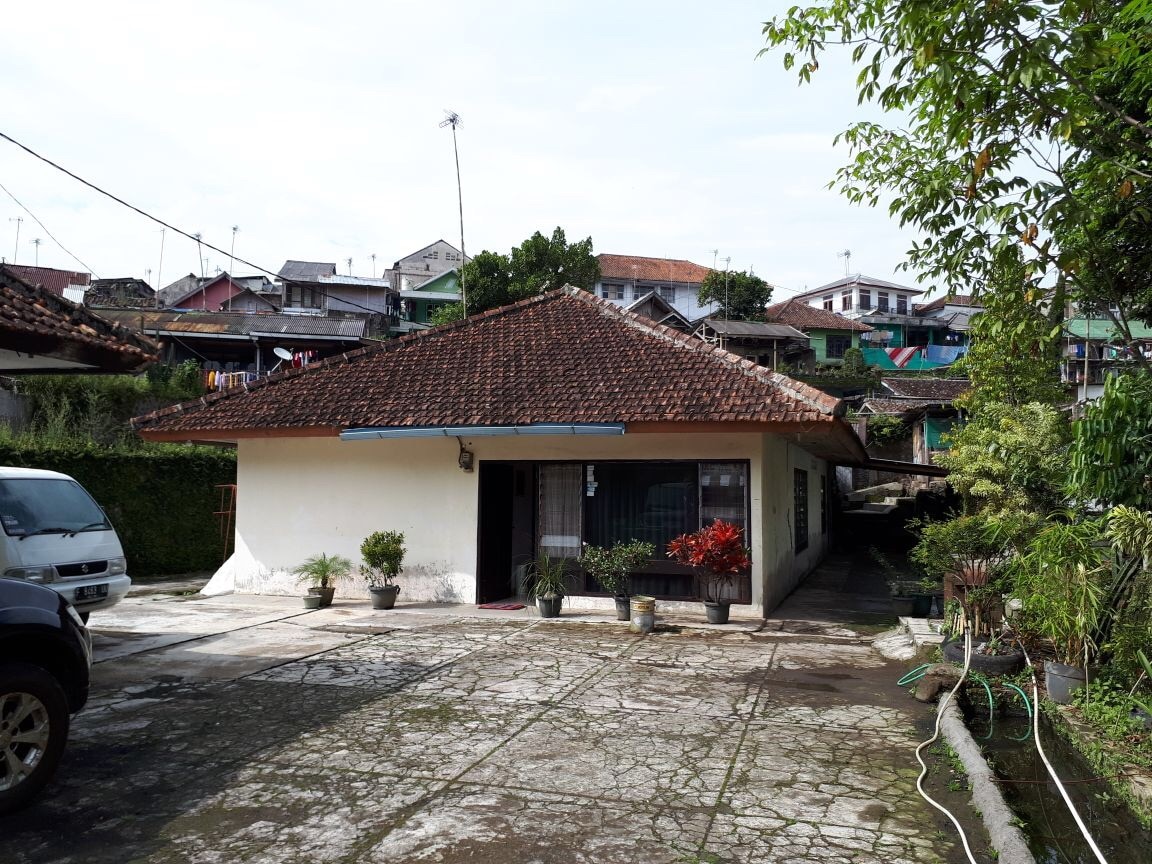 A single-story house is viewed from the front, surrounded by a court paved with concrete. The roof features traditional tiles, and pots with greenery are placed near the entrance. Nearby structures are visible in the background, set against a bright sky.