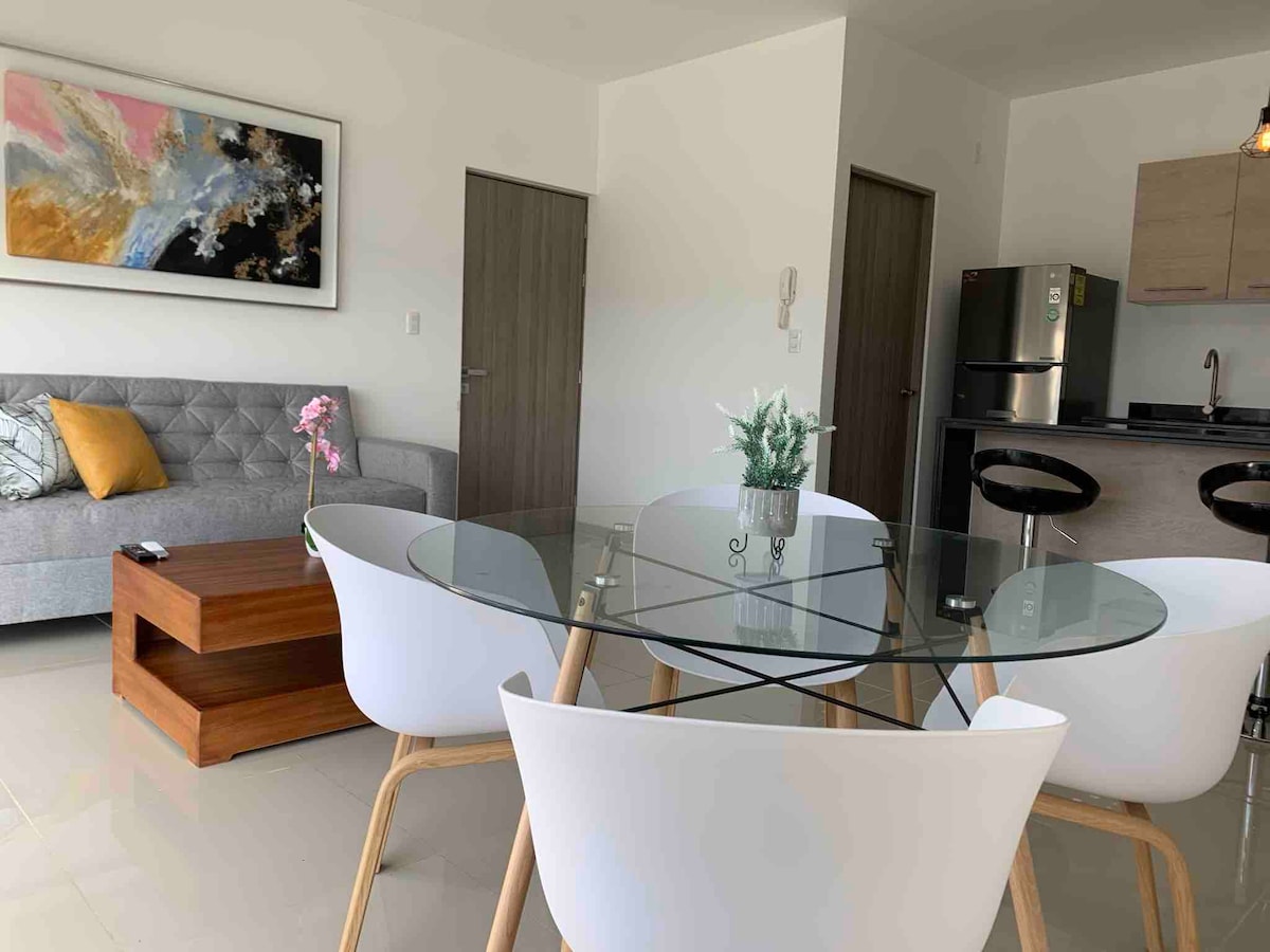 A modern dining area features a glass table surrounded by four white chairs. A stylish wooden coffee table sits nearby. A gray sofa with a decorative pillow is positioned against the wall, while a contemporary artwork hangs above.