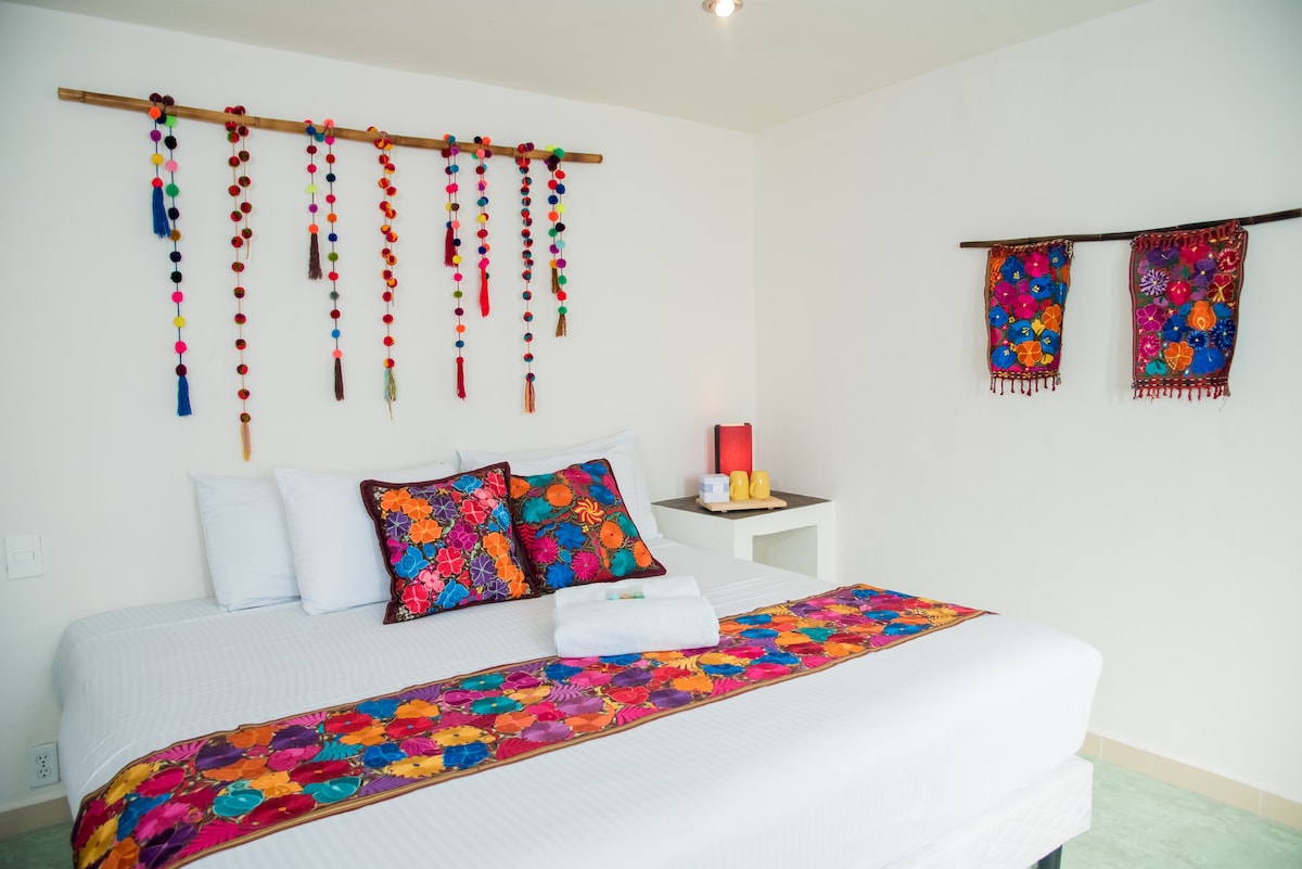 A spacious room features a comfortable bed adorned with colorful accent pillows and a decorative bed runner. Handcrafted wall hangings, including vibrant textiles, add a touch of local art to the white walls, while a small side table holds essentials.