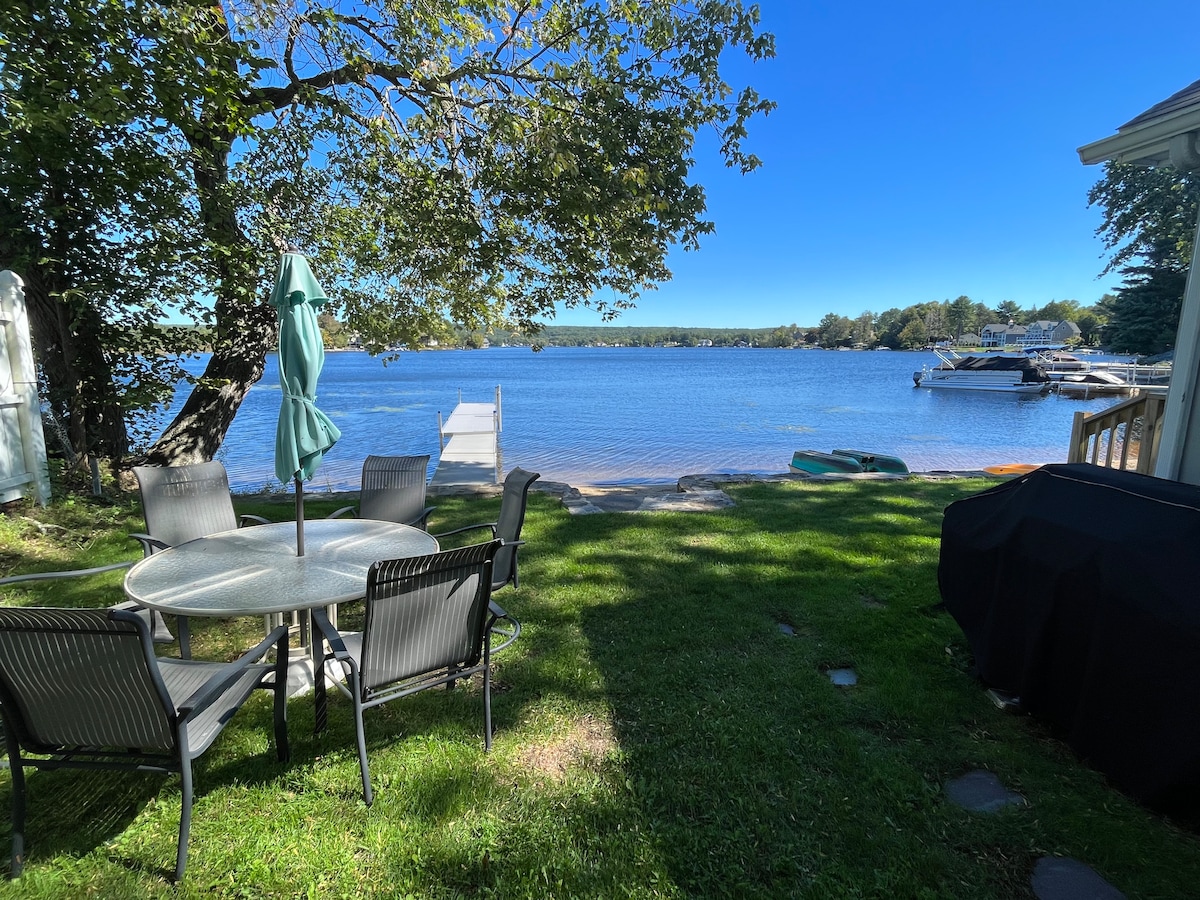 A green lawn extends towards the tranquil waterfront, featuring a round outdoor dining table surrounded by six chairs. A grill is positioned nearby, while a paddleboat rests on the shoreline. Trees provide shade, with clear blue water visible in the background.