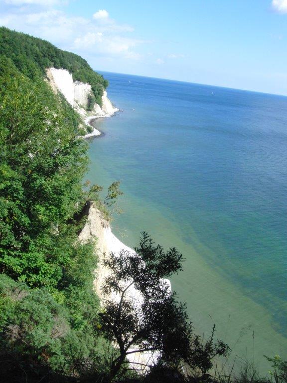 The image captures a coastal view featuring white cliffs along the shoreline. Dense green trees frame the left side, while calm blue waters extend towards the horizon. A clear sky with scattered clouds enhances the natural beauty of the landscape.