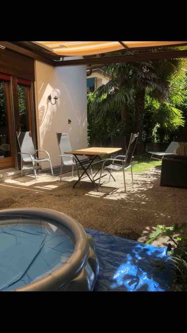 A private garden area features a small table with two chairs and two lounge chairs surrounded by greenery. A shaded seating area is visible, with a hint of a hot tub partially seen on the left, providing an inviting outdoor space.