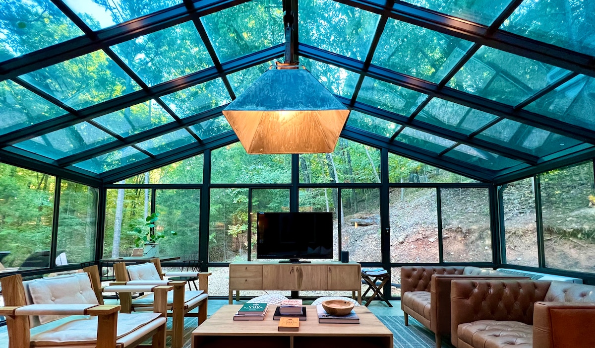 A spacious glass room is surrounded by lush greenery, featuring comfortable seating arrangements, including a wood accent chair and a leather sofa. A large flat-screen TV is mounted on the wall, complemented by a wooden console table. Soft lighting is provided by a distinctive pendant lamp, enhancing the inviting atmosphere.