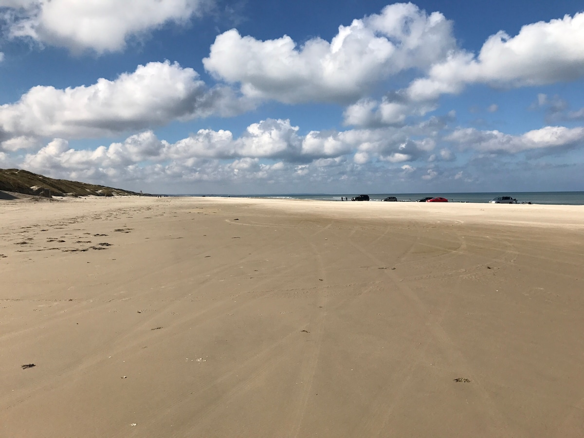 A vast sandy beach stretches towards the horizon, marked by gentle waves lapping at the shore. Numerous clouds create a textured sky above, casting scattered shadows on the sand. In the distance, a few cars are parked, blending into the serene coastal landscape.